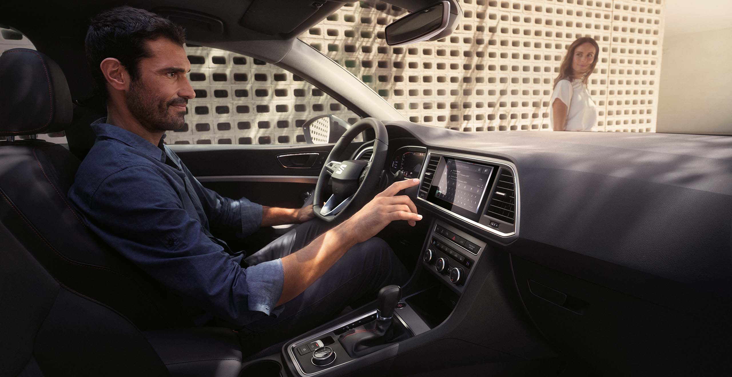 Man driving a SEAT Atec 2024 company car focused on the road, using infotainment system, while a woman stands in the background by a patterned wall, looking at him